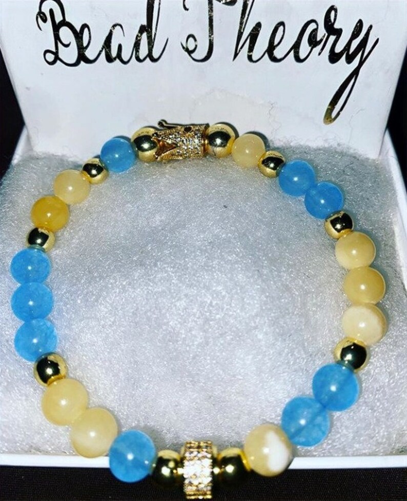 Puede incluir: Un brazalete de cuentas azules y amarillas con detalles dorados y un dije de corona. El brazalete est&aacute; en una caja blanca con las palabras "Bead Theory" impresas en la parte superior.