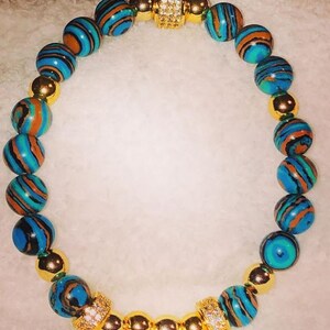 Puede incluir: Un brazalete de cuentas con estampado azul y marr&oacute;n con detalles dorados y un cierre dorado con diamantes de imitaci&oacute;n transparentes.