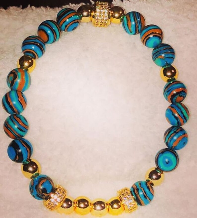 Puede incluir: Un brazalete de cuentas con estampado azul y marr&oacute;n con detalles dorados y un cierre dorado con diamantes de imitaci&oacute;n transparentes.