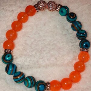 Puede incluir: Un brazalete con cuentas naranjas y azules. Las cuentas est&aacute;n separadas por separadores dorados con diamantes de imitaci&oacute;n transparentes. El brazalete est&aacute; hecho con un cord&oacute;n el&aacute;stico.