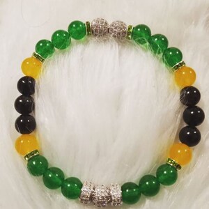 Puede incluir: Un brazalete de cuentas verdes, amarillas y negras con detalles de strass plateados y transparentes. El brazalete tiene dos detalles plateados y de strass en cada extremo.