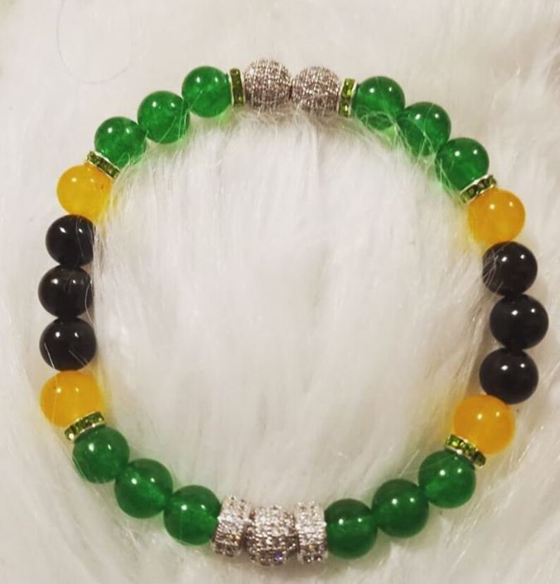 Puede incluir: Un brazalete de cuentas verdes, amarillas y negras con detalles de strass plateados y transparentes. El brazalete tiene dos detalles plateados y de strass en cada extremo.
