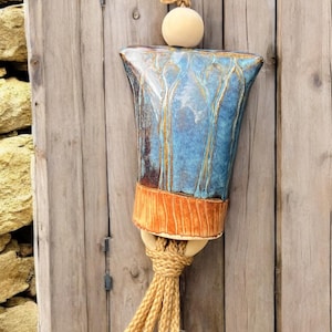 May include: A handcrafted ceramic wall hanging with a blue and brown glazed finish. The piece features a unique shape, a tan rope, and a decorative tassel. The wall art is approximately 12 inches in height.