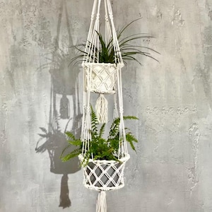 Op de afbeelding: Een macramé plantenhouder met twee niveaus, gemaakt van wit geweven touw. De bovenste mand bevat een groene plant met lange, dunne bladeren, en de onderste mand een varen. De houder hangt tegen een grijze, getextureerde muur.