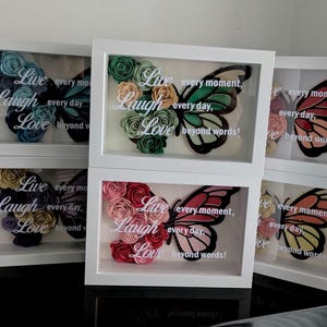 May include: White framed art pieces with butterfly designs. Each butterfly is made of colourful paper flowers and a printed wing. The text "Live, Laugh, Love" is displayed on each piece, along with the phrase "every moment, every day, beyond words!"