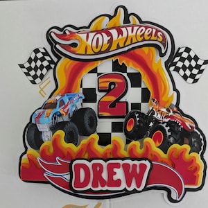 May include: A colorful Hot Wheels cake topper featuring two monster trucks, flames, and the name "DREW." The number "2" is prominently displayed, with checkered flags on either side. The Hot Wheels logo is at the top.