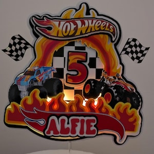 May include: A custom Hot Wheels-themed birthday cake topper featuring two monster trucks, flames, a checkered flag, and the name "ALFIE". The number "5" is prominently displayed, with the Hot Wheels logo at the top.