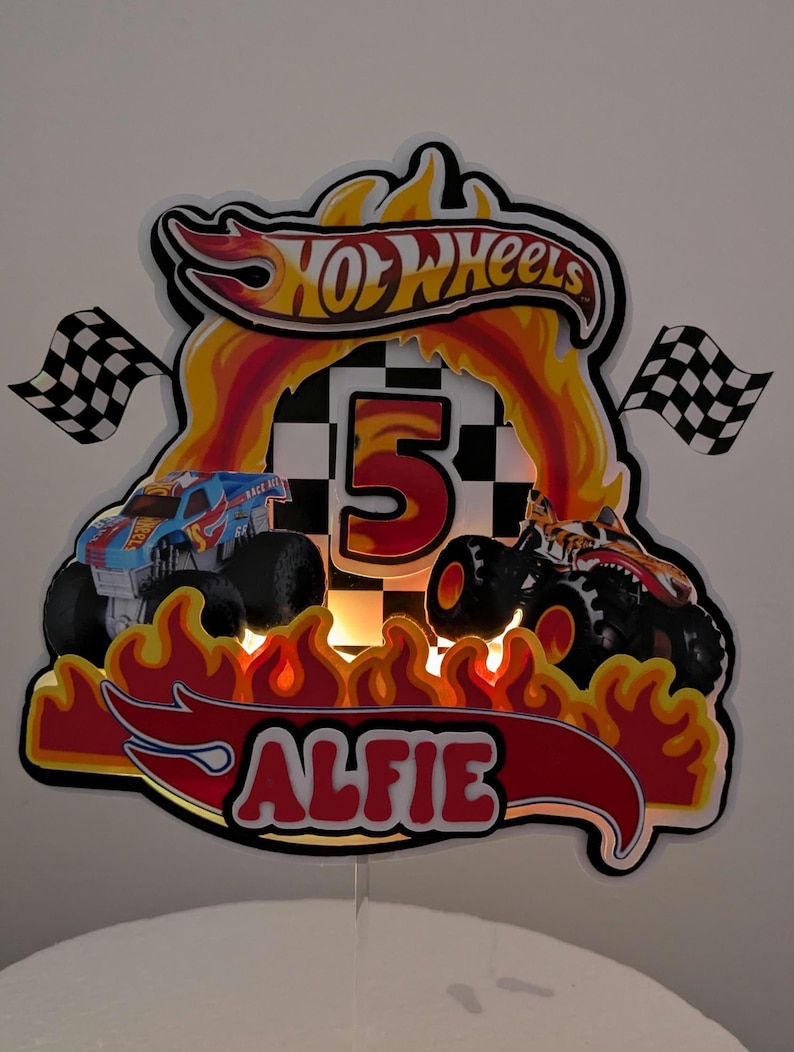 May include: A custom Hot Wheels-themed birthday cake topper featuring two monster trucks, flames, a checkered flag, and the name "ALFIE". The number "5" is prominently displayed, with the Hot Wheels logo at the top.