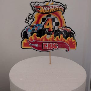 May include: A Hot Wheels-themed cake topper featuring a red banner with the name "ERIC", a large number "4", and the Hot Wheels logo. The design includes flames and monster trucks, perfect for a birthday celebration.