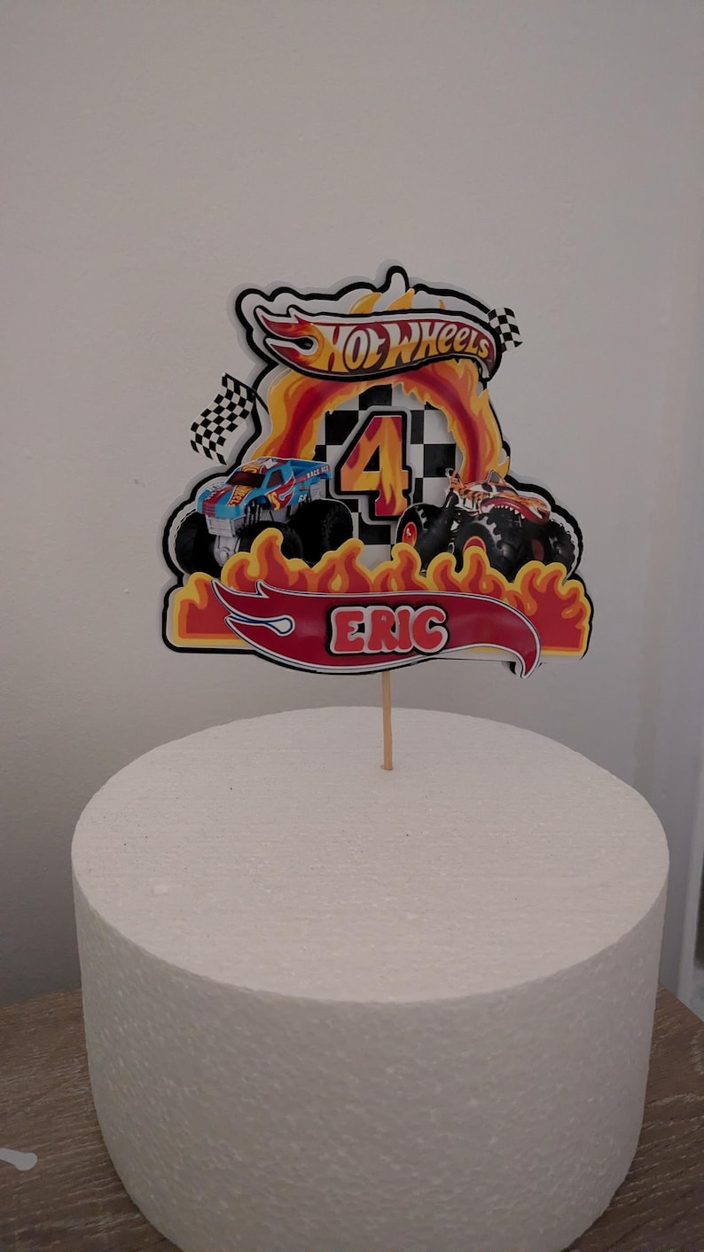 May include: A Hot Wheels-themed cake topper featuring a red banner with the name "ERIC", a large number "4", and the Hot Wheels logo. The design includes flames and monster trucks, perfect for a birthday celebration.