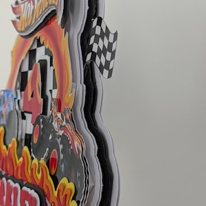 May include: A layered Hot Wheels-themed cake topper with a racing theme. The design includes the Hot Wheels logo, flames, a monster truck, a checkered flag, and the number 4. The name "Oakley" is also visible.