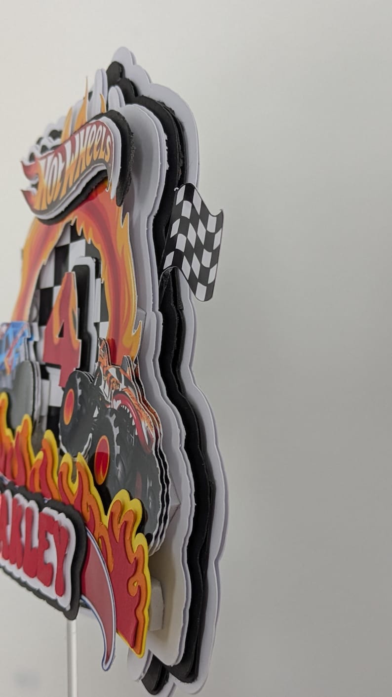 May include: A layered Hot Wheels-themed cake topper with a racing theme. The design includes the Hot Wheels logo, flames, a monster truck, a checkered flag, and the number 4. The name "Oakley" is also visible.