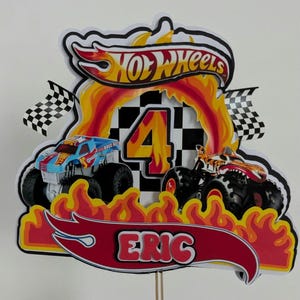May include: A Hot Wheels-themed birthday cake topper featuring two monster trucks, flames, and the name "Eric." The number "4" is prominently displayed, along with the Hot Wheels logo and checkered flags. The design is predominantly red, yellow, and black.