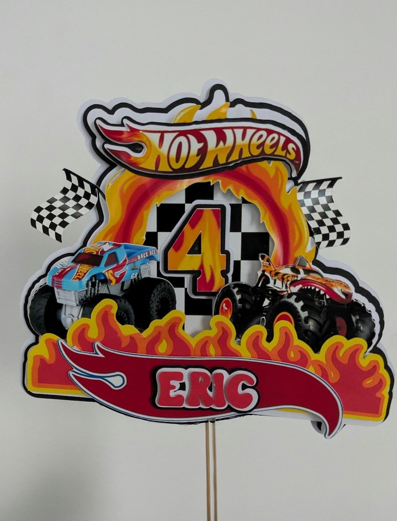 May include: A Hot Wheels-themed birthday cake topper featuring two monster trucks, flames, and the name "Eric." The number "4" is prominently displayed, along with the Hot Wheels logo and checkered flags. The design is predominantly red, yellow, and black.