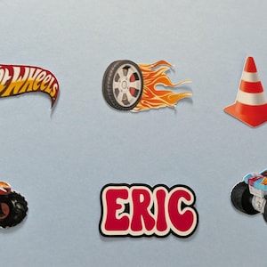 May include: A collection of colorful Hot Wheels-themed stickers. Includes the Hot Wheels logo, a tire with flames, a traffic cone, two monster trucks, and a sticker with the name "ERIC" in red and white.