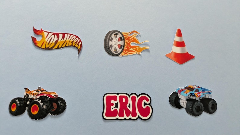 May include: A collection of colorful Hot Wheels-themed stickers. Includes the Hot Wheels logo, a tire with flames, a traffic cone, two monster trucks, and a sticker with the name "ERIC" in red and white.
