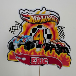May include: A colorful Hot Wheels-themed cake topper featuring two monster trucks, flames, and the name "Eric." The number "4" is prominently displayed, along with the Hot Wheels logo and checkered flags. The topper is mounted on a wooden stick.