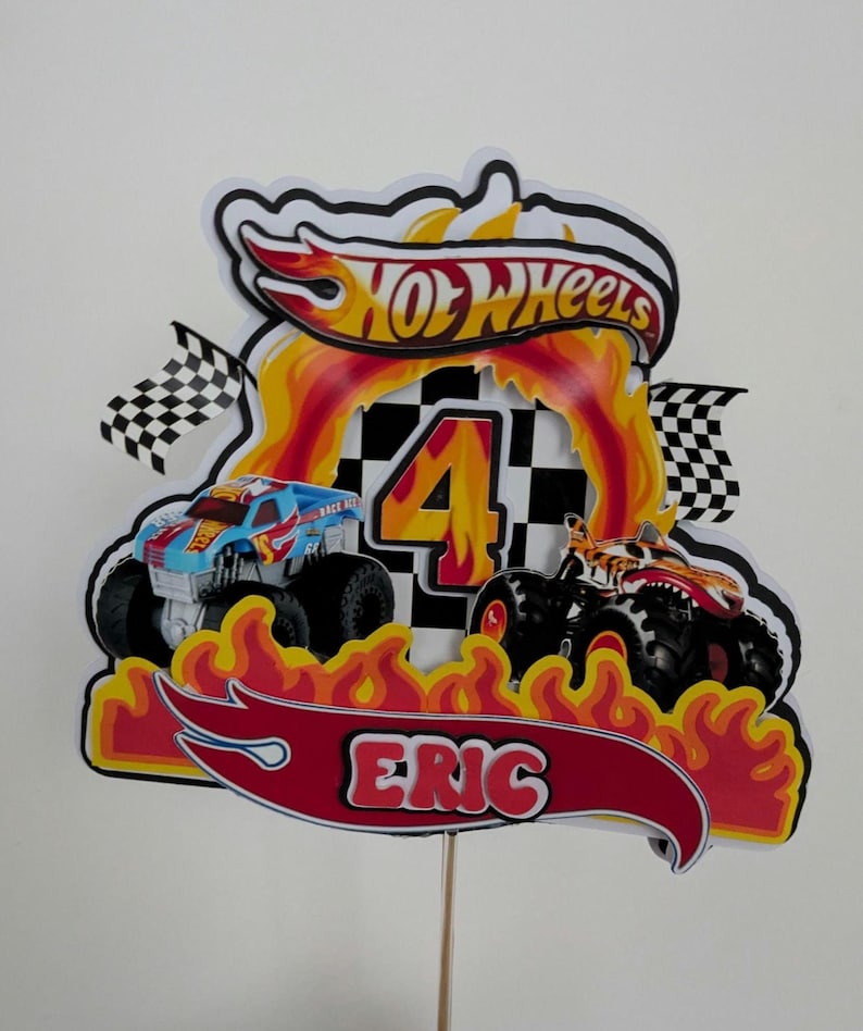 May include: A colorful Hot Wheels-themed cake topper featuring two monster trucks, flames, and the name "Eric." The number "4" is prominently displayed, along with the Hot Wheels logo and checkered flags. The topper is mounted on a wooden stick.