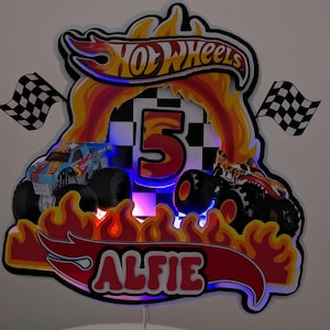 May include: A colorful Hot Wheels-themed birthday cake topper. The design features two monster trucks, flames, checkered flags, and the number 5. The name "ALFIE" is displayed in red lettering. The Hot Wheels logo is at the top.