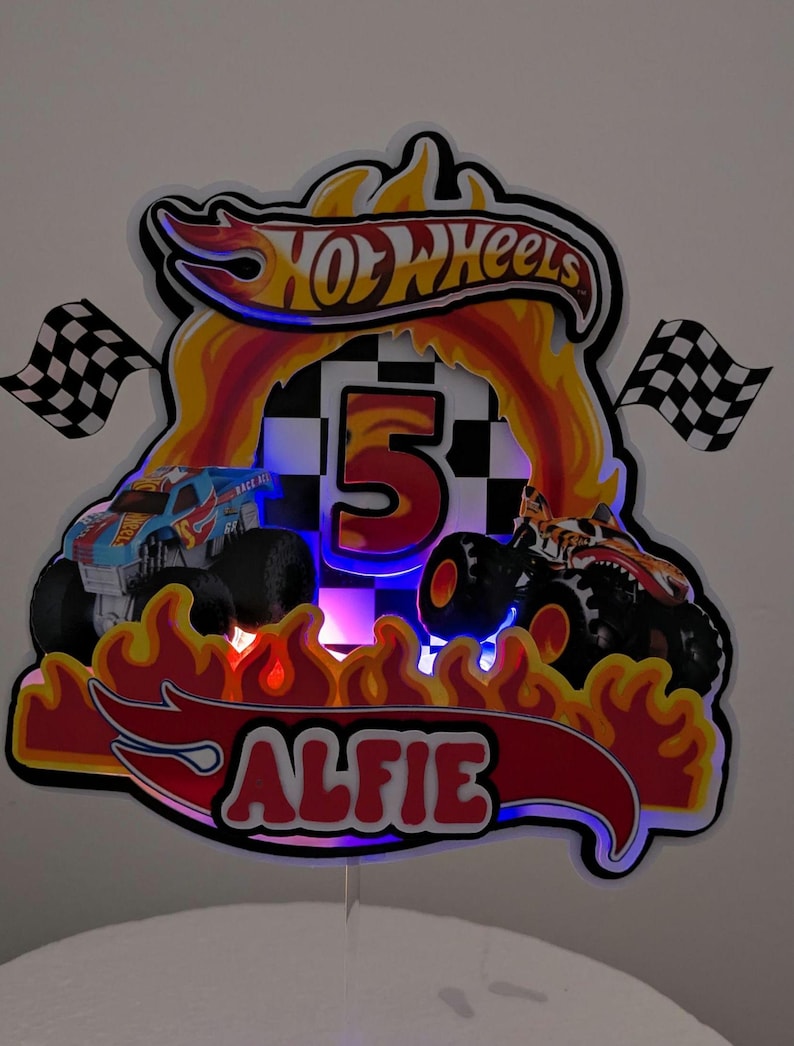 May include: A colorful Hot Wheels-themed birthday cake topper. The design features two monster trucks, flames, checkered flags, and the number 5. The name "ALFIE" is displayed in red lettering. The Hot Wheels logo is at the top.
