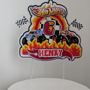 May include: Cake topper featuring a Hot Wheels design with two monster trucks, flames, and the number 6. The name "HENRY" is displayed in red. Includes two checkered flags. The topper is on a white cake.