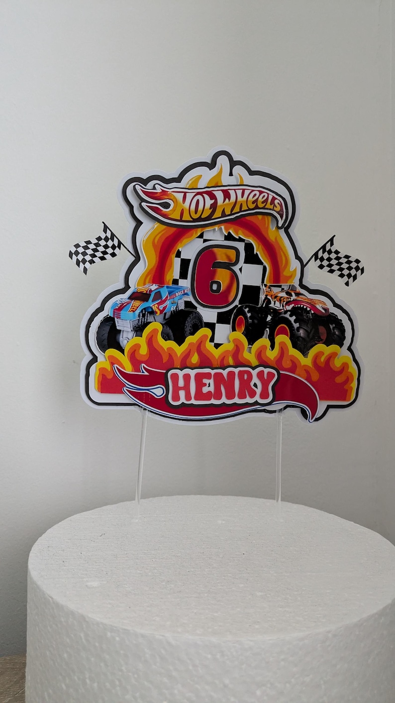 May include: Cake topper featuring a Hot Wheels design with two monster trucks, flames, and the number 6. The name "HENRY" is displayed in red. Includes two checkered flags. The topper is on a white cake.