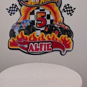 May include: Cake topper featuring the Hot Wheels logo, flames, and two monster trucks. The number 5 is in the center, with the name "Alfie" below. Two checkered flags flank the design. The topper is on a white cake.