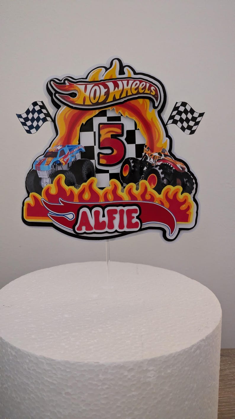 May include: Cake topper featuring the Hot Wheels logo, flames, and two monster trucks. The number 5 is in the center, with the name "Alfie" below. Two checkered flags flank the design. The topper is on a white cake.