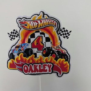 May include: A colorful Hot Wheels-themed cake topper featuring a red and yellow flame design. The topper includes the Hot Wheels logo, a checkered flag, the number 4, and the name "OAKLEY" in a red and white font. Two toy monster trucks are also featured.