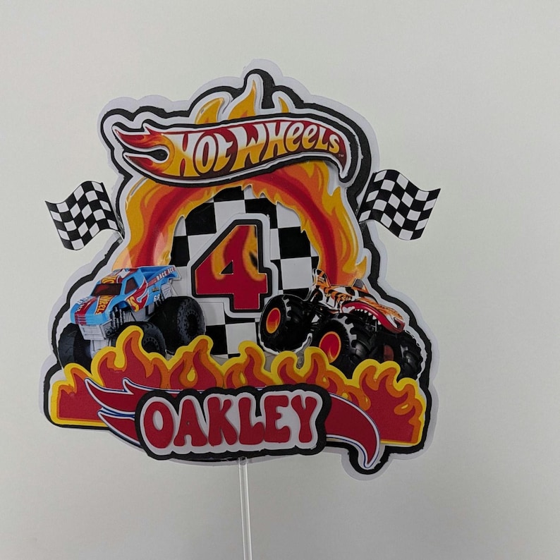 May include: A colorful Hot Wheels-themed cake topper featuring a red and yellow flame design. The topper includes the Hot Wheels logo, a checkered flag, the number 4, and the name "OAKLEY" in a red and white font. Two toy monster trucks are also featured.