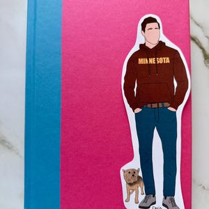 May include: A hardcover book with a pink cover and a blue spine. A sticker of a person in a brown hoodie with "MINNESOTA" printed on it, blue jeans, and gray shoes is on the cover. A small dog sticker is also on the cover.