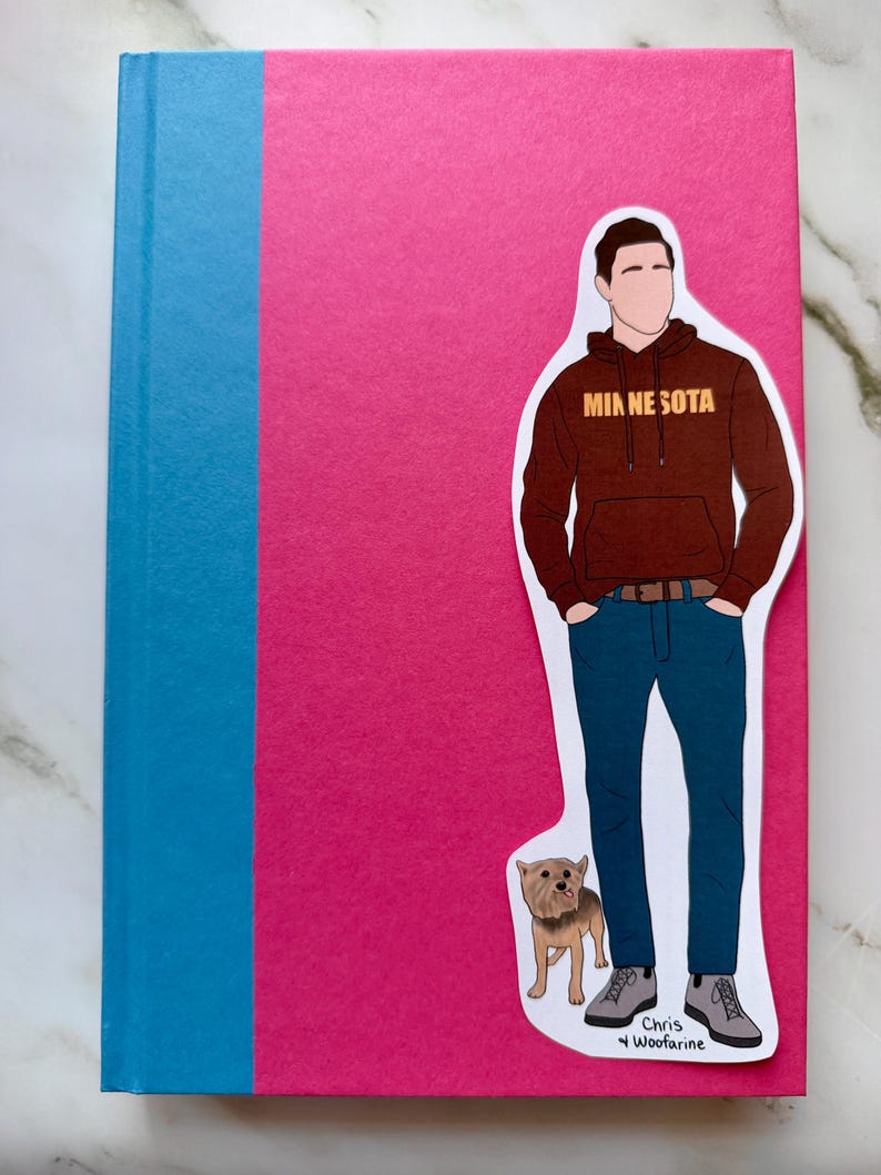 May include: A hardcover book with a pink cover and a blue spine. A sticker of a person in a brown hoodie with "MINNESOTA" printed on it, blue jeans, and gray shoes is on the cover. A small dog sticker is also on the cover.