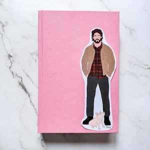 May include: A pink journal with a sticker of a man in a brown jacket and plaid shirt, with a pig. The sticker is placed on the front cover of the journal. The journal is rectangular and has a smooth texture.
