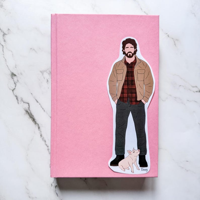 May include: A pink journal with a sticker of a man in a brown jacket and plaid shirt, with a pig. The sticker is placed on the front cover of the journal. The journal is rectangular and has a smooth texture.