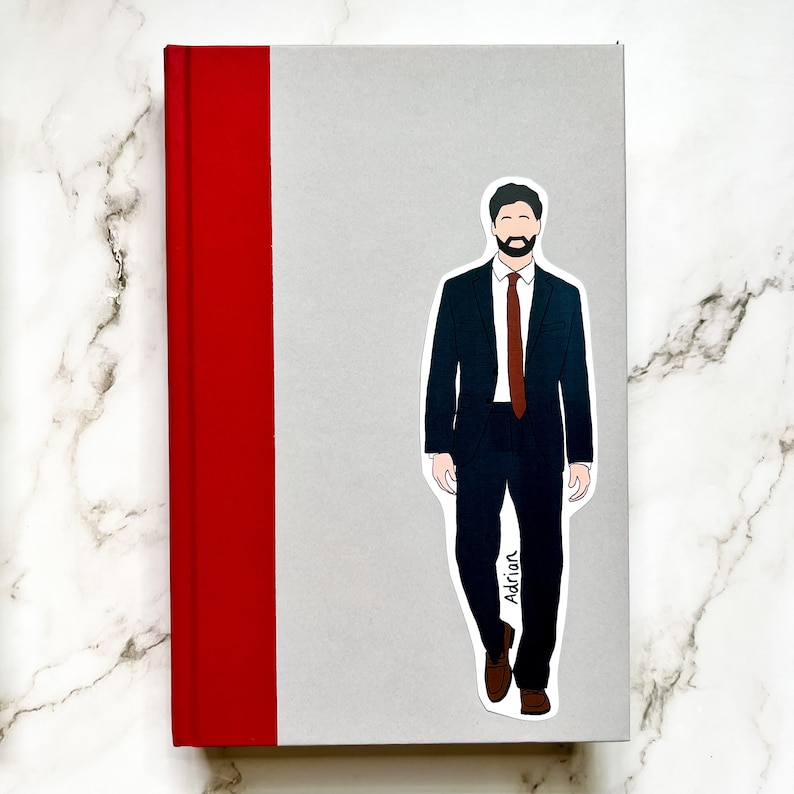 May include: A sticker of a man in a dark suit and brown tie, with the name "Adrian" printed on the bottom right. The sticker is placed on a gray book with a red spine.