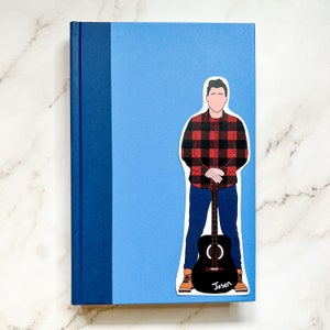 May include: A light blue journal with a dark blue spine. A sticker of a person in a red and black plaid shirt, blue jeans, and brown boots holding a black guitar is on the front. The name "Jason" is written on the guitar.