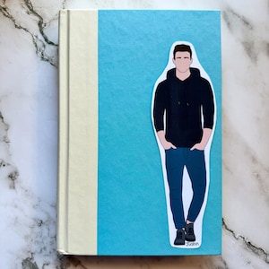 May include: A hardcover book with a light blue cover and a cream-colored spine. A sticker of a person in a black hoodie and blue jeans is on the cover. The sticker has the name "Justin" at the bottom.