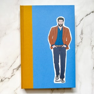 May include: A hardcover book with a blue cover and a mustard yellow spine. A sticker of a man in a brown blazer, blue shirt, and jeans is on the cover. The sticker has the name "Daniel" written on it.