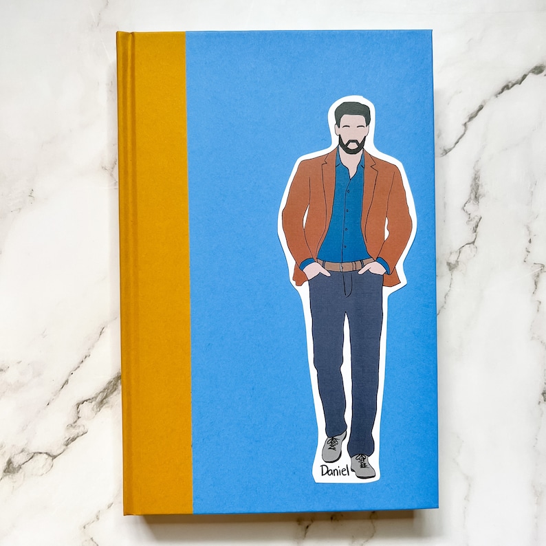 May include: A hardcover book with a blue cover and a mustard yellow spine. A sticker of a man in a brown blazer, blue shirt, and jeans is on the cover. The sticker has the name "Daniel" written on it.