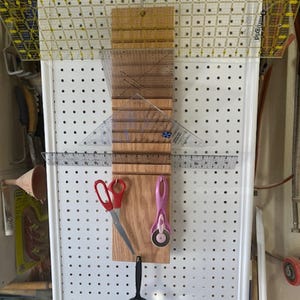 May include: A wooden board with a ruler, a pair of scissors, a rotary cutter, and a magnifying glass hanging on a pegboard.