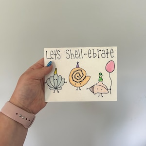 Op de afbeelding: Een handgetekende wenskaart met de tekst "Let's Shell-ebrate" boven drie cartoon schelpen met feesthoedjes en een roze ballon. De schelpen zijn in lichtblauw, oranje en roze.