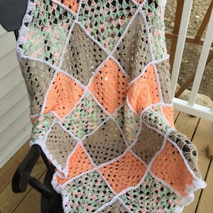 May include: A crocheted blanket with a patchwork design in peach, green, and brown. The blanket is edged with white crochet and is draped over a wooden rocking chair.