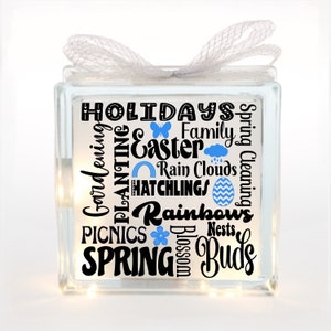 May include: A glass block with a white ribbon and a string of fairy lights. The block features a design of words and images associated with spring, including "Holidays", "Easter", "Rain Clouds", "Hatchlings", "Rainbows", "Spring Cleaning", "Picnics", "Spring", and "Buds".