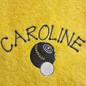 May include: A bright yellow towel featuring the embroidered name CAROLINE in black thread, positioned above a black lawn bowl with a white jack ball, showcasing a textured fabric surface with detailed stitching throughout the design.
