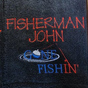 May include: Two black rectangular towels featuring embroidered text and graphics. The left towel reads SNOTBOX and the right reads FISHERMAN JOHN in red. Both feature a blue and white fishing bobber graphic with the text GONE FISHIN' in blue.