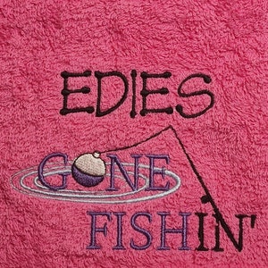 May include: A bright pink terry cloth towel featuring black and purple embroidery. The text reads EDIES GONE FISHIN with a graphic of a fishing rod and a purple and white bobber floating in water ripples, all stitched onto the fabric surface.