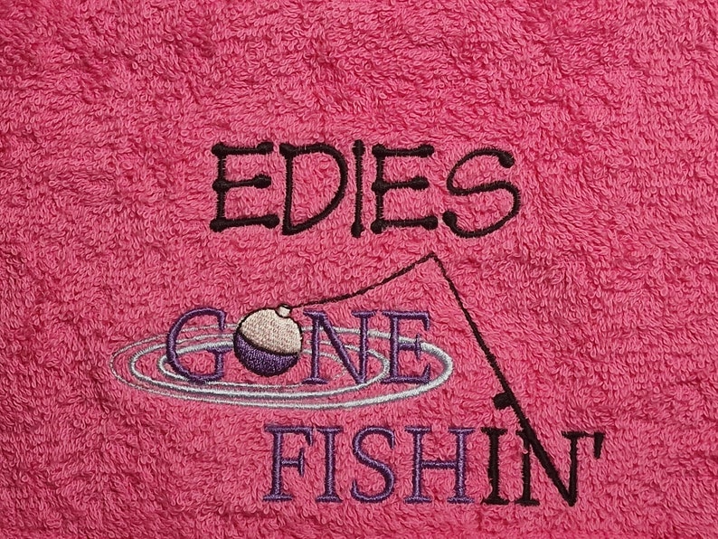May include: A bright pink terry cloth towel featuring black and purple embroidery. The text reads EDIES GONE FISHIN with a graphic of a fishing rod and a purple and white bobber floating in water ripples, all stitched onto the fabric surface.