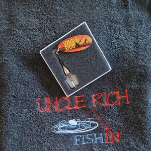 May include: A dark gray towel embroidered with the red text UNCLE RICH and the blue text GONE FISHIN with a fishing rod graphic. A square box contains a silver fishing lure with an oval orange tag reading UNCLE RICH.