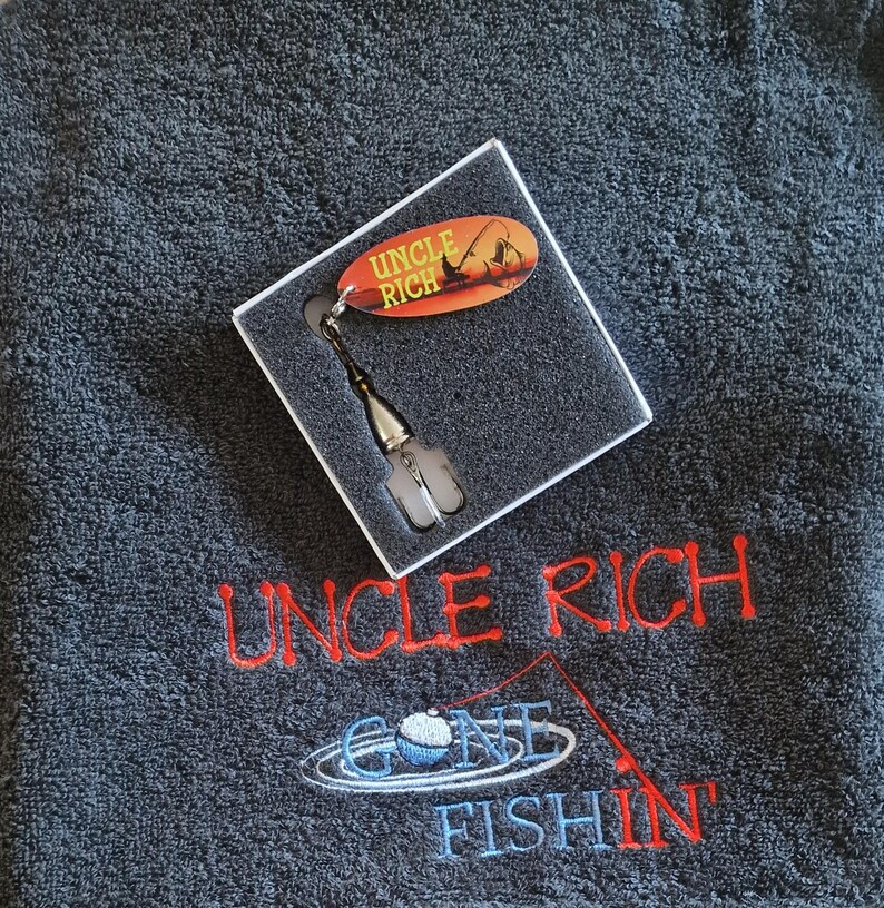 May include: A dark gray towel embroidered with the red text UNCLE RICH and the blue text GONE FISHIN with a fishing rod graphic. A square box contains a silver fishing lure with an oval orange tag reading UNCLE RICH.
