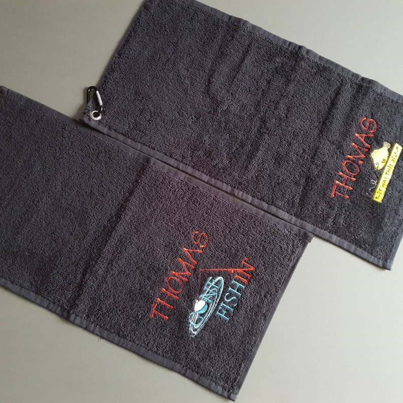 May include: Two dark gray terry cloth towels featuring embroidery. One towel displays the text "THOMAS FISHIN" with a fishing bobber graphic. The other towel features the name "THOMAS" and a yellow fish graphic with the text "IT WAS THIS BIG".